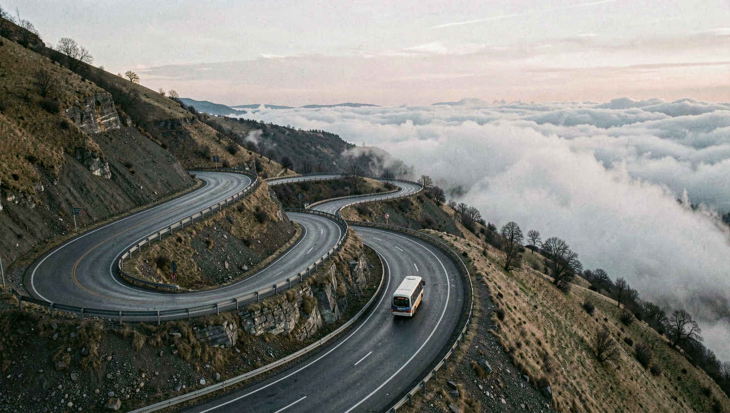 Passstrasse mit gestapelten Serpentinen an einem kahlen Felshang, tief liegende Wolken zwischen den Kehren, ein einzelnes Postauto in einer Kurve, kaltes Licht kurz nach Sonnenaufgang, Panoramaaufnahme von oben